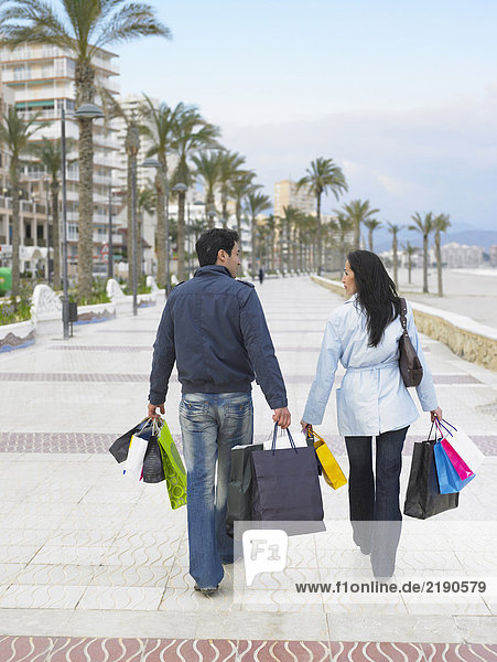 Rückansicht eines Paares  das mit Einkaufstaschen entlang eines palmengesäumten Bürgersteigs geht. Alicante  Spanien.
