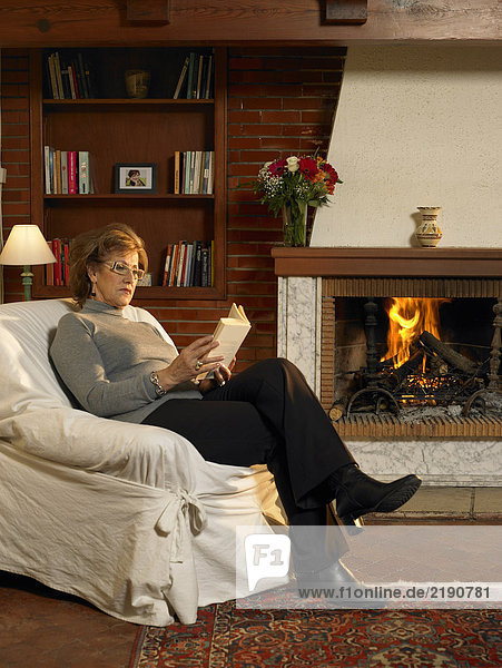 Senior woman reading book by fire in living room