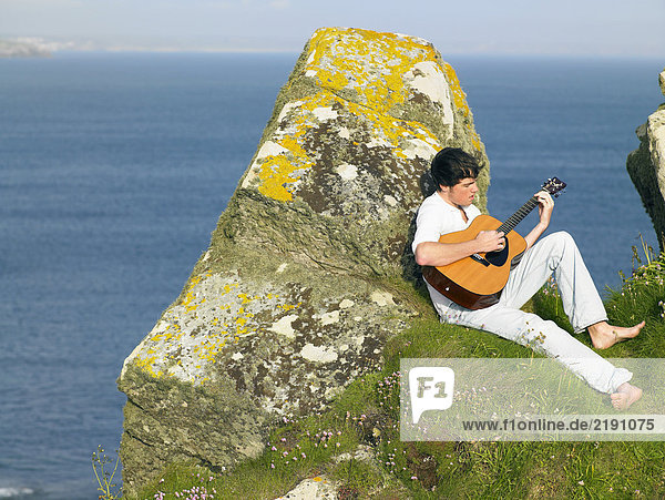 Junger Mann sitzt an einer Klippe und spielt Gitarre.