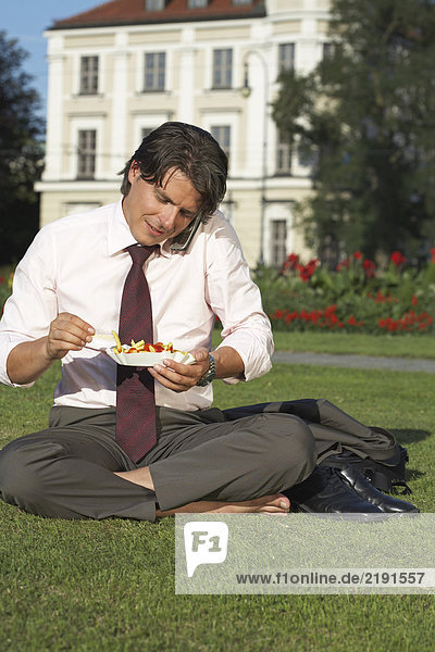 Businessman on square sitting in meadow on the mobile whilst eating chips laughing.