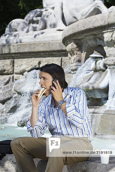 Ein Geschäftsmann sitzt am Rand des Brunnens und isst ein Sandwich  während er telefoniert.