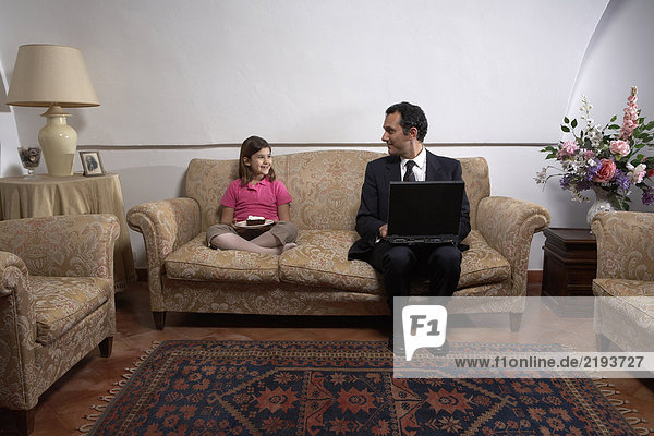 Man using laptop looking at daughter (6-8) on sofa with cake