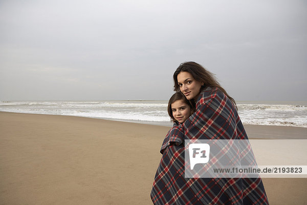 Mother and daughter (9-11) wrapped in blanket on beach