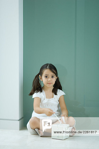 Kleines Mädchen sitzt auf dem Boden  bindet das Band an das Geschenk  schaut in die Kamera.
