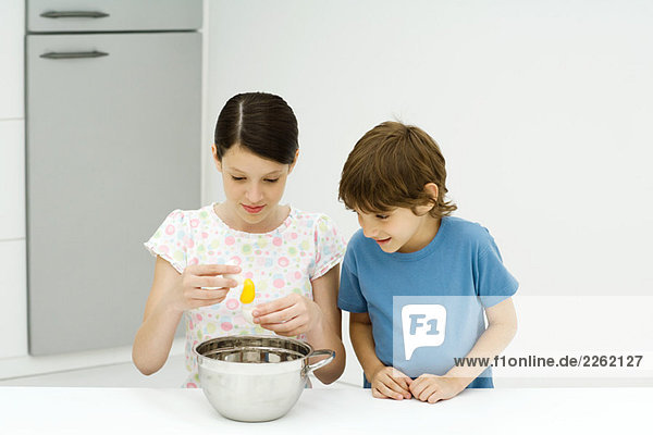 Zwei junge Geschwister kochen zusammen,  Mädchen knackt Ei,  beide schauen nach unten.