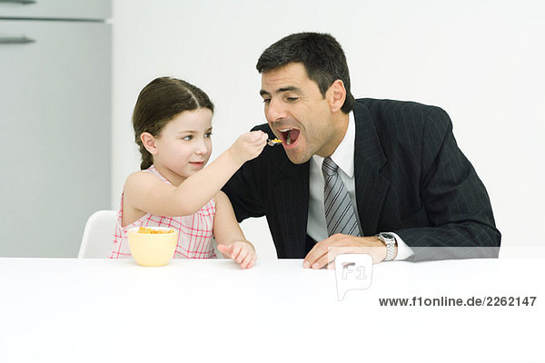 Kleines Mädchen füttert ihren Vater mit Getreide  beide sitzen am Tisch.