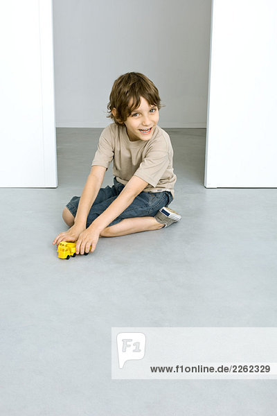 Kleiner Junge sitzt auf dem Boden  spielt mit Spielzeugwagen  lächelt in die Kamera.