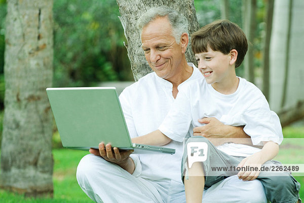 Mature man holding grandson on lap  both looking at laptop computer and smiling
