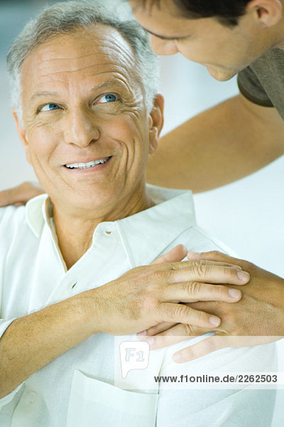 Mature man and adult son smiling at each other  son's hand on father's shoulder  cropped view