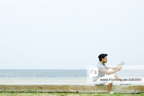 Mann auf niedriger Wand am Strand sitzend  Lesebuch  Seitenansicht