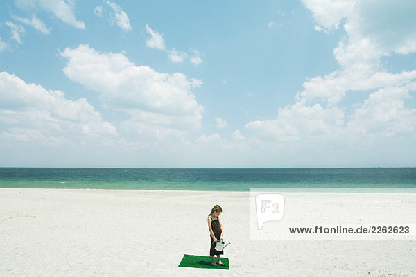 Mädchen steht auf Kunstrasenplatz am Strand  hält Gießkanne  Blick in den hohen Winkel
