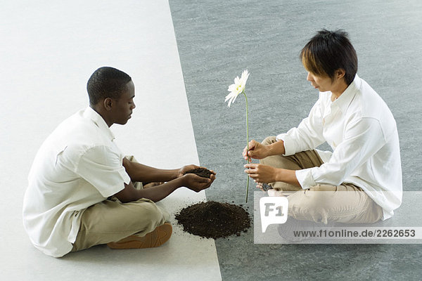 Zwei Männer sitzen von Angesicht zu Angesicht auf dem Boden  der eine hält eine Handvoll Erde  der andere eine Blume aus.