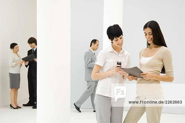 Zwei Geschäftsfrauen stehen zusammen  schauen auf die Mappe  eine hält Handy  Mitarbeiter im Hintergrund