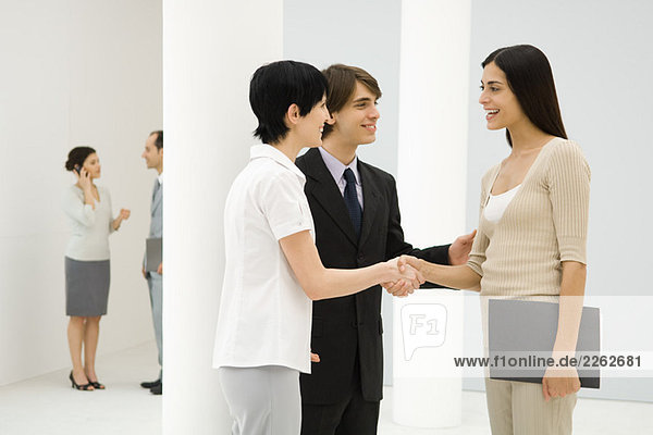 Zwei Geschäftsfrauen stehen neben einem männlichen Kollegen  schütteln sich die Hände und lächeln sich an.