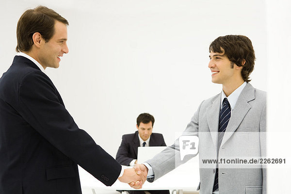 Zwei männliche Profis  die sich die Hände schütteln  lächeln sich an  ein Kollege sitzt im Hintergrund.