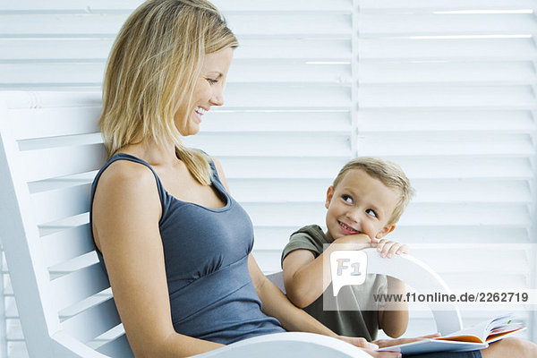 Woman sitting in chair next to son  holding book  both smiling at each other