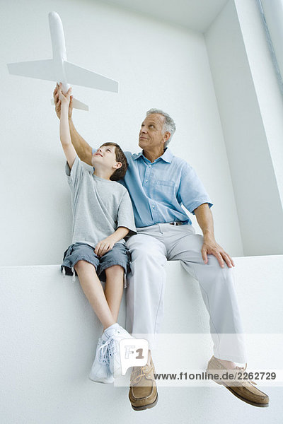 Grandfather and grandson sitting side by side  holding up toy airplane together