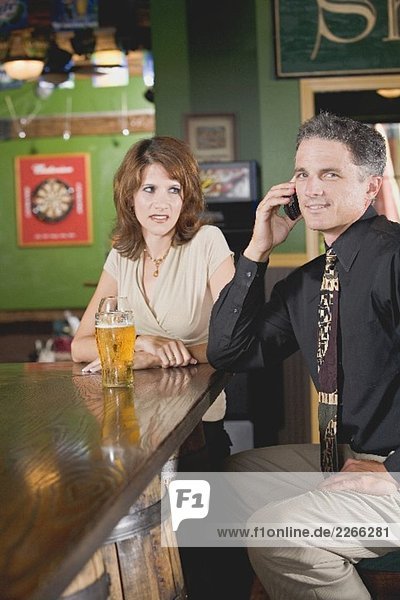 Woman standing disapprovingly beside man on phone in a pub