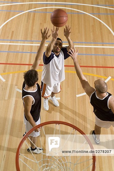 Ein Basketballspieler schießt den Basketball  während zwei weitere Spieler versuchen  seinen Schuss zu blockieren.