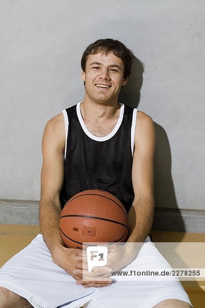 Ein junger Mann sitzt auf einer Bank mit Armen um einen Basketball.