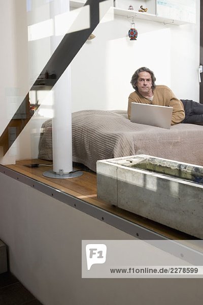 Ein Mann  der mit einem Laptop auf seinem Bett liegt.