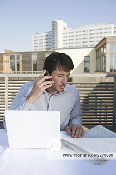 Ein Geschäftsmann sitzt auf einer Dachterrasse mit Zeitung  Handy und Laptop.