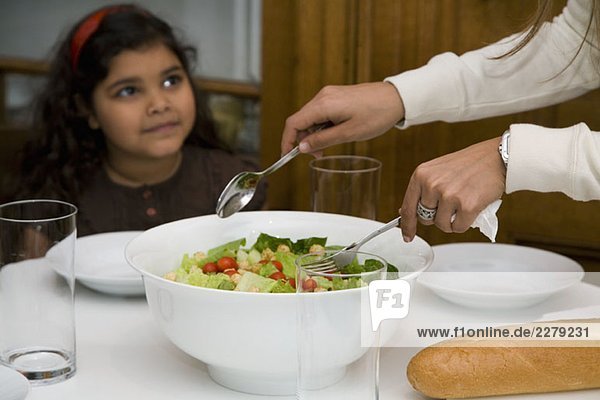 Ein Mädchen sitzt am Tisch und eine Frau serviert Salat.