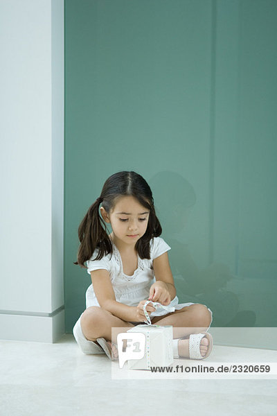 Kleines Mädchen sitzt auf dem Boden und bindet ein Band an ein Geschenk.