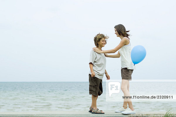Geschwister stehen von Angesicht zu Angesicht am Strand  Mädchenhände auf den Schultern des Bruders  Junge hält Ballon