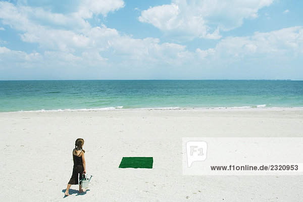Girl at the beach walking toward patch of artificial turf  carrying watering can  rear view