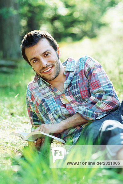 Man sitting on the ground outdoors  holding book  smiling at camera