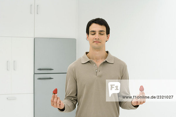Mann steht in der Küche mit geschlossenen Augen und hält Erdbeere in jeder Hand.