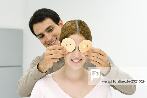 Mann  der hinter einer Frau steht und ihre Augen mit Keksen bedeckt  beide lächelnd.