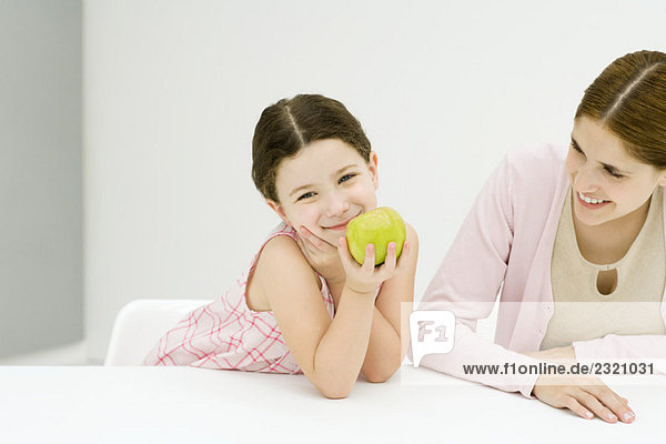 Kleines Mädchen am Tisch sitzend mit Mutter  Hand unter dem Kinn  Apfel haltend  Kamera lächelnd