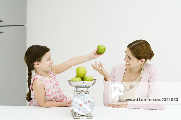 Mutter und Tochter wiegen Äpfel auf der Waage  lächeln sich an  Mädchen hält einen Apfel hoch.