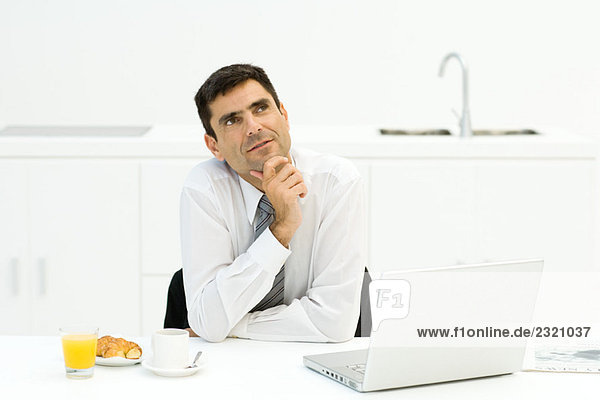 Geschäftsmann am Tisch sitzend mit Laptop und Frühstück  Hand unter dem Kinn  aufblickend