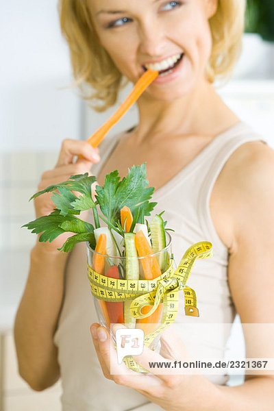 Woman biting into carrot  holding glass full of vegetables tied with measuring tape