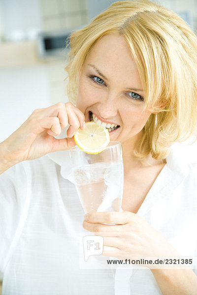 Frau hält ein Glas Wasser  beißt in die Zitronenscheibe  schaut in die Kamera