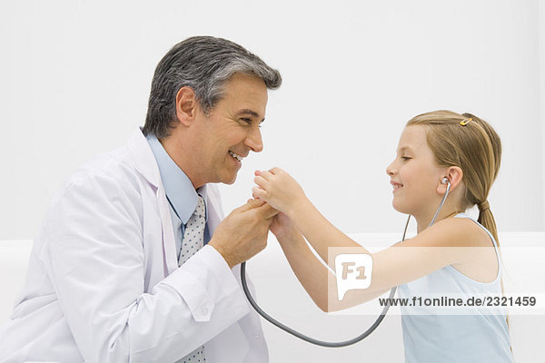 Der Arzt spricht ins Stethoskop  während das kleine Mädchen lächelt.