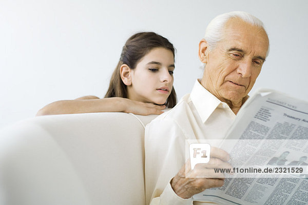 Preteen Mädchen schaut Großvater über die Schulter  während er Zeitung liest.