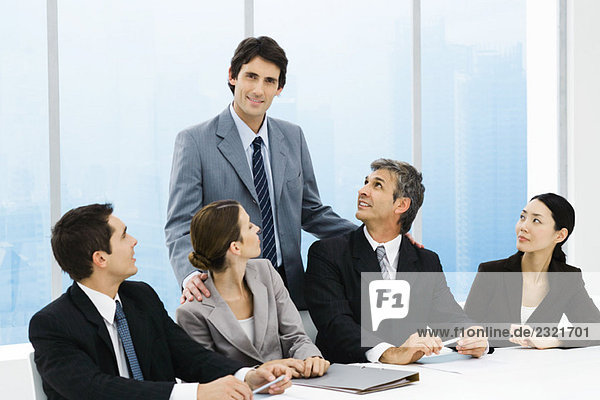 Geschäftsleiter steht hinter einer Gruppe von Kollegen am Besprechungstisch und lächelt in die Kamera.