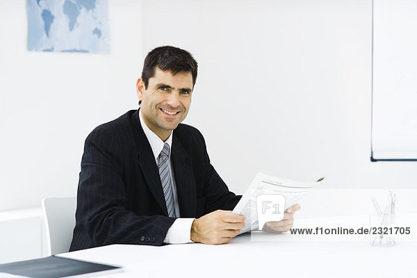 Geschäftsmann am Schreibtisch im Büro  Zeitung lesen  Kamera lächeln