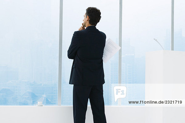 Businessman standing by window  holding document under arm  thinking  rear view