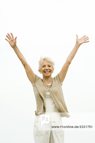 Seniorin steht draußen mit erhobenen Armen in der Luft und lächelt die Kamera an.