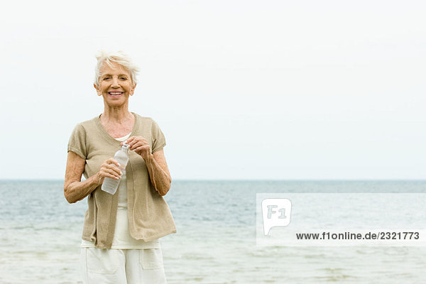 Seniorin steht am Strand  hält eine Flasche Wasser  lächelt die Kamera an.