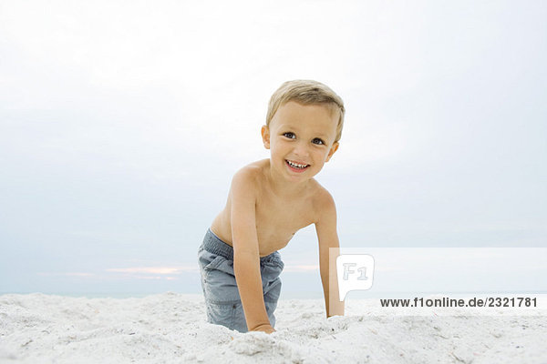Kleiner Junge kauernd am Strand  lächelnd vor der Kamera  Porträt