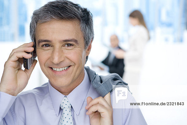 Mature businessman using cell phone in office  holding jacket over shoulder  smiling at camera