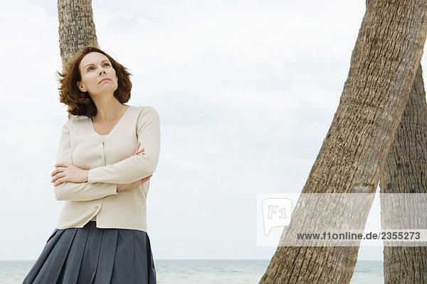 Mature woman leaning against palm tree with arms folded  looking up