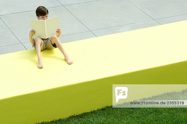 Boy sitting on low wall outdoors  reading book  high angle view