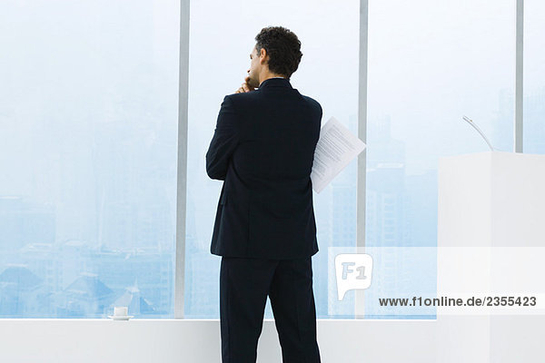 Businessman standing in front of window  looking out  holding document  rear view